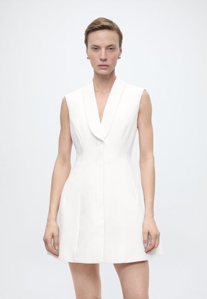 Kvinne med kort hår iført en ermeløs, hvit skreddersydd kjole med slag og gullfargede øreringer, stående foran en ensfarget bakgrunn.