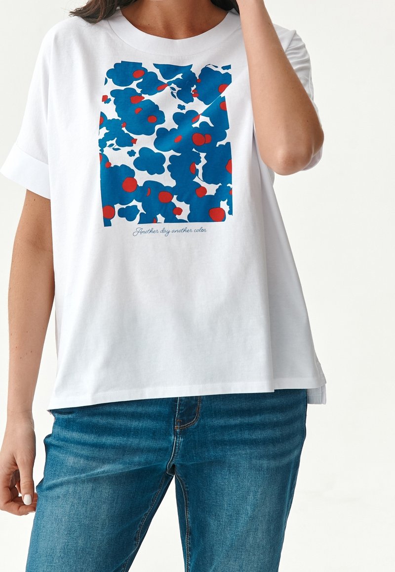 Chemisette blanche avec imprimé floral carré bleu et rouge et texte "Un jour de plus, une couleur de plus", portée avec un jean bleu.