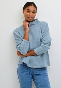 Pullover in maglia azzurro chiaro, morbido, con colletto alto, spalline scese e polsini a costine. Indossato con jeans aderenti azzurri chiari.