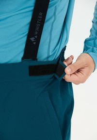Person passt den Klettverschluss an einer dunkelgrünen Hose mit schwarzen Whistler-Trägern an und trägt ein hellblaues Langarmshirt.
