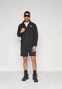 Sort udendørsjakke med fuld lynlås og logo, kombineret med sorte shorts. Sorte vandrestøvler og mørke solbriller fuldender looket.