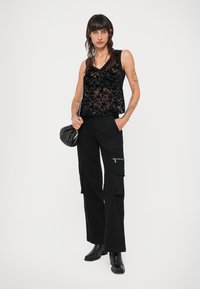 Zwart doorzichtig tanktop met een bloemenkantpatroon, gecombineerd met zwarte cargobroek met zakken. Model draagt zwarte enkellaarsjes en houdt een kleine tas vast.