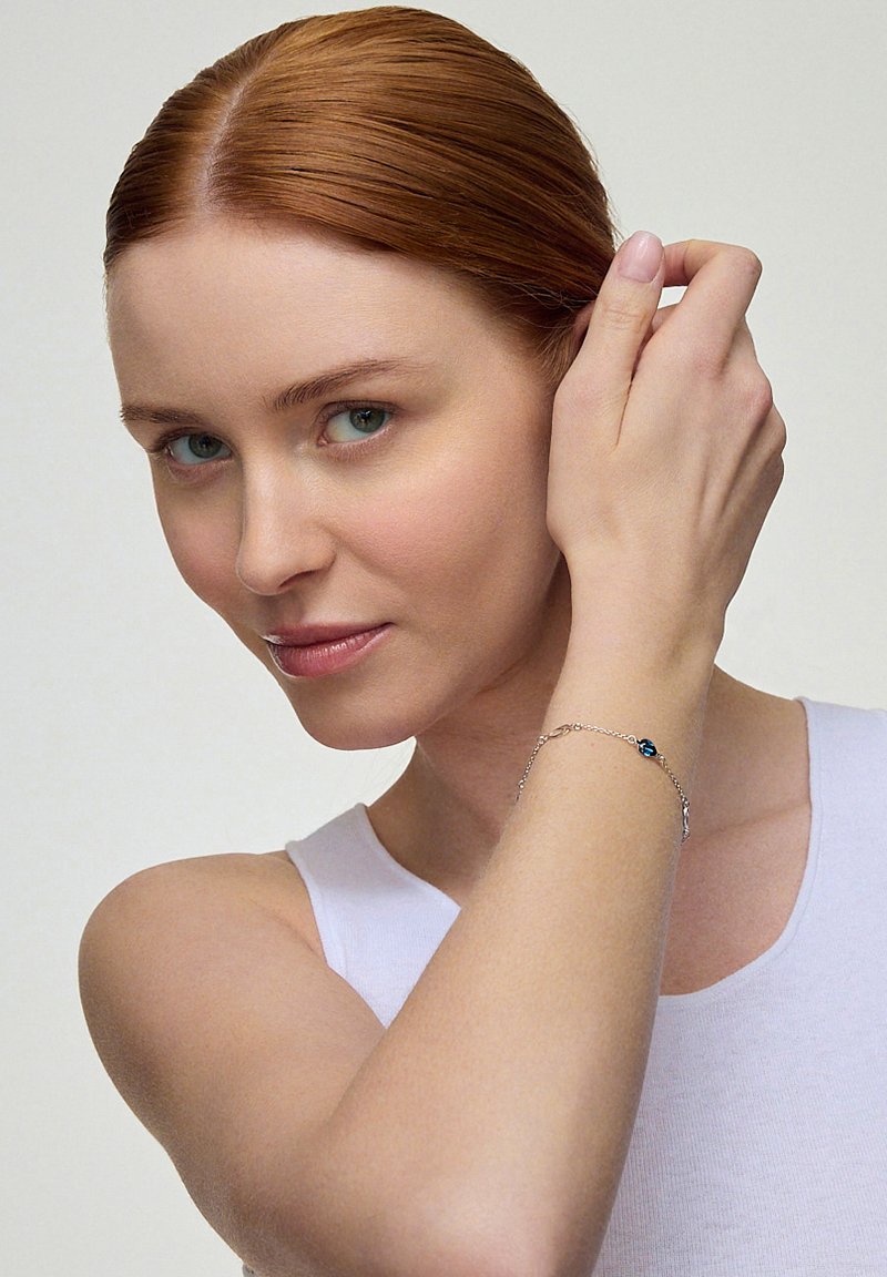 Srebrna bransoletka z akcentem niebieskiego kamienia, z delikatnym wzorem łańcuszka. Noszona na jasnej skórze, na minimalistycznym tle.
