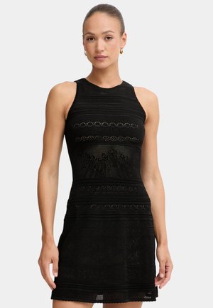 Donna che indossa un vestito nero lavorato a maglia senza maniche con strisce orizzontali decorate, in piedi davanti a uno sfondo chiaro e uniforme.