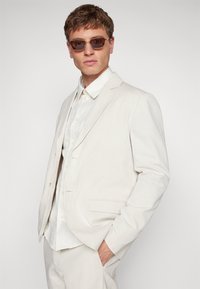 Lys beige dressjakke med to frontlommer, sammen med en teksturert, hvit skjorte. Modellen har på seg brune rektangulære solbriller.