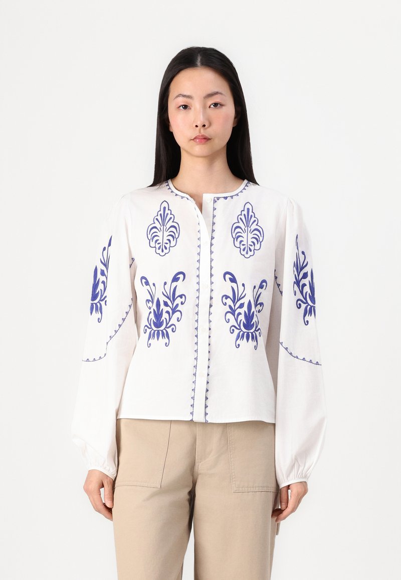 Blouse en coton blanc avec broderie florale bleue, encolure ronde, fermeture à boutons à l'avant et manches longues et amples. Couture texturée le long des bords.