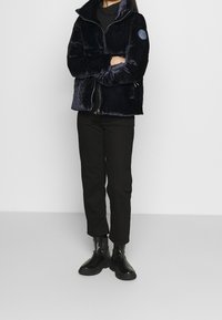 Giacca in pile blu navy con zip e colletto alto, caratterizzata da un patch rotondo sulla manica, abbinata a pantaloni neri e stivali neri lucidi.