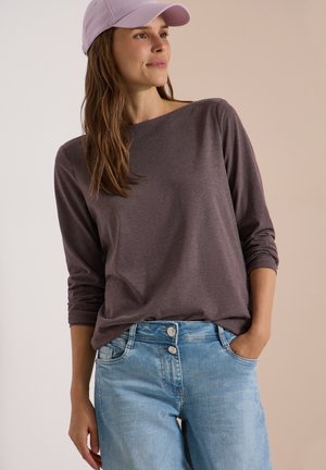 Femme portant une casquette lavande, un haut gris foncé à manches longues, un jean bleu clair taille haute avec deux boutons, main dans la poche, regard vers la droite.