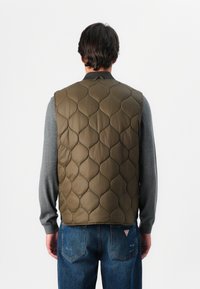 Olive quilted vest z wysokim kołnierzem, o gładkiej fakturze i subtelnym wzorze w diamenty, noszony na szarej bluzie z długim rękawem.