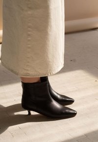 Svarta läderankelboots med spetsig tå och låg klack, kombinerade med en beige, knälång, strukturerad kjol. Diskret glans på bootsen.