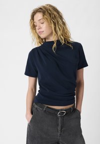 Top navy a maniche corte con design drappeggiato, tessuto morbido, colletto a giro, e vestibilità rilassata, abbinato a pantaloni in denim grigi a vita alta e cintura nera.