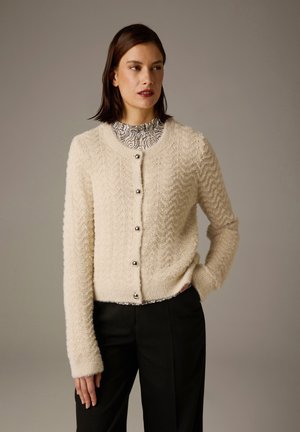 Kremfarbener, strukturierter Cardigan mit Zickzackmuster, versehen mit runden silbernen Knöpfen und einem weichen, flauschigen Material. Lange Ärmel und ein runder Halsausschnitt.