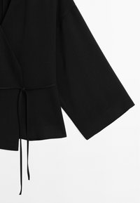 Top nero avvolgente con maniche larghe, realizzato in tessuto morbido. Presenta un legaccio in vita e un orlo peplum, che offre una vestibilità comoda.