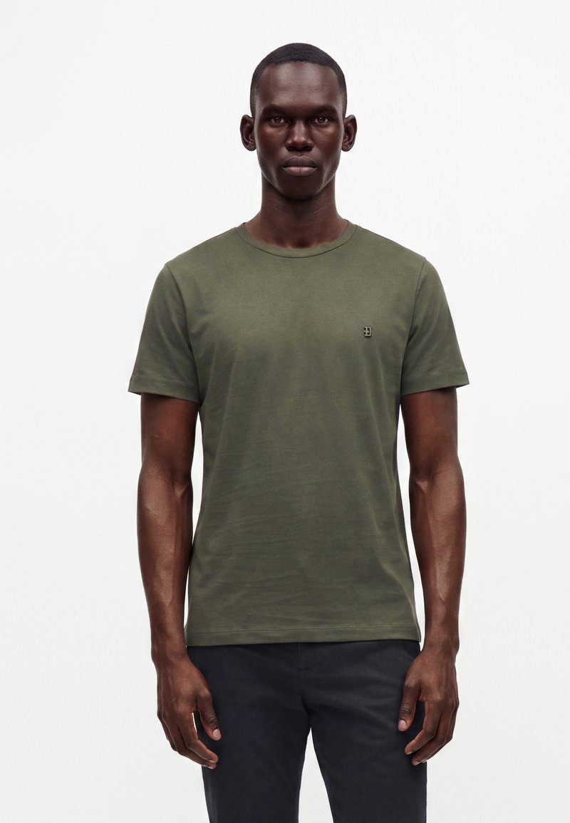 Homme à la peau foncée portant un t-shirt vert olive à manches courtes et un pantalon noir, debout devant un fond blanc uni.