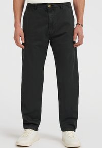Mand iført sorte lige bukser og hvide sneakers, stående med hænderne afslappet langs siden mod en hvid baggrund.