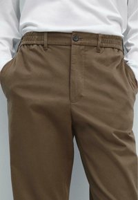 Bruine broeken met een elastische tailleband en een knoopsluiting. Voorzien van zijzakken en een zachte textuur.