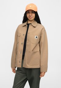 Γυναίκα που φοράει μπεζ τζάκετ με κουμπιά και τρεις μεγάλες τσέπες, μαύρο πουκάμισο, χακί παντελόνι και πορτοκαλί καπέλο μπέιζμπολ, στέκεται μπροστά σε ένα απλό φόντο.