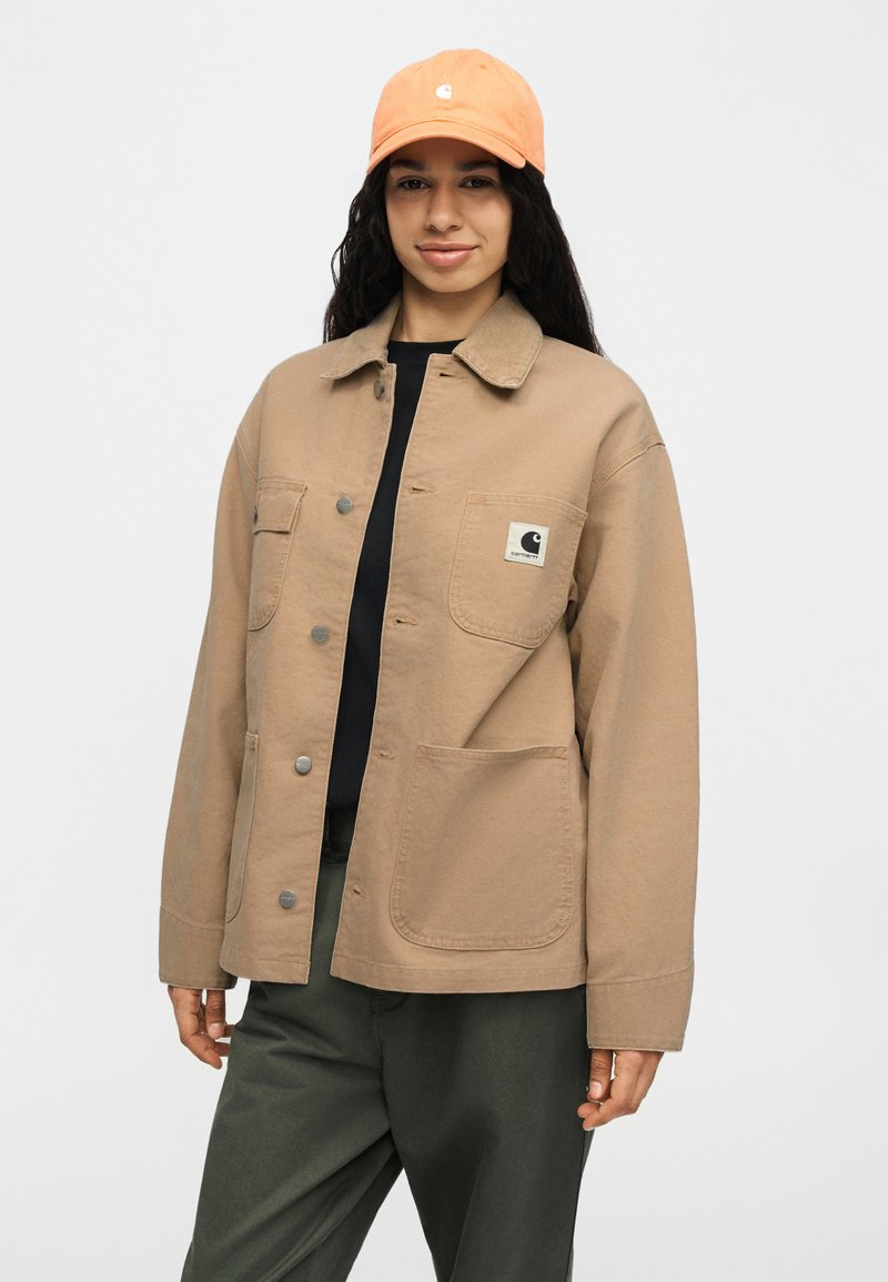 Vrouw met een zandkleurig jasje met knopen en drie grote zakken, zwart shirt, olijfgroene broek en een oranje baseballpet, staande tegen een effen achtergrond.