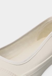 Gros plan sur une chaussure décontractée sans lacets de couleur blanc cassé, à texture lisse et avec des coutures le long du bord d'ouverture.
