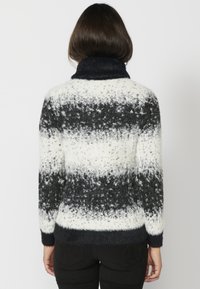 Frau in einem flauschigen schwarz-weiß gestreiften Rollkragenspullover, von hinten betrachtet, kombiniert mit schwarzen Hosen.