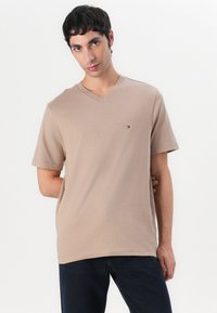 T-shirt beige à col en V en mélange de coton, avec des manches courtes et un petit logo brodé sur la poitrine gauche. Texture lisse.