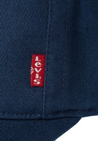Blå denimstoff med et synlig rødt Levi's-merke sydd på venstre side. Stoffet har et twill-vev og rene linjer.