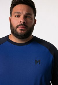 Blå skjorta med marinblå accenter, rund halsringning och korta raglansärmar. Har en svart logotyp "M" på bröstet. Slät tygkvalitet.