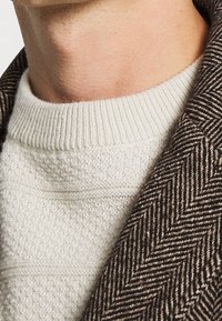 Primo piano del collo di una persona che indossa un maglione a maglia testurizzato color crema e un cappotto a motivo herringbone marrone.