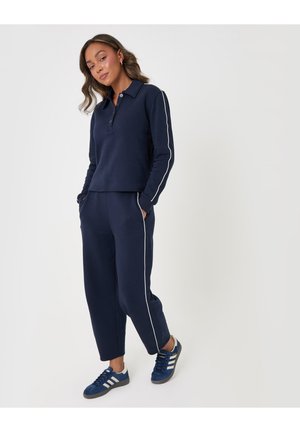 Mujer con chándal azul marino con franjas laterales blancas y zapatillas azules, de pie con las manos en los bolsillos frente a un fondo liso.