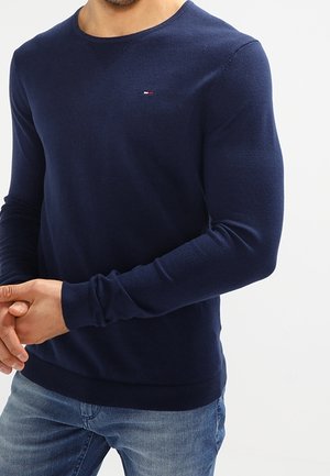 Mann trägt einen enganliegenden marineblauen Pullover mit langen Ärmeln, einem kleinen roten, weißen und blauen Logo auf der Brust und hellblaue Jeans.