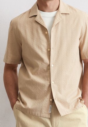 Hombre con camisa beige de manga corta y botones sobre camiseta blanca con las manos en los bolsillos contra un fondo liso.