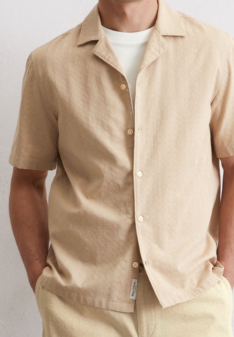 Homme portant une chemise beige à manches courtes boutonnée sur un t-shirt blanc, les mains dans les poches, sur un fond uni.