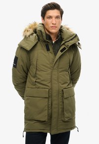Olivengrønt parka med faux fur-indfatning på hætten, lynlås foran og store frontlommer. Har justerbar bund og detaljer med sort patch.