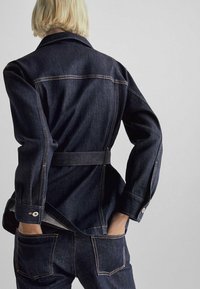 Ciemna kurtka dżinsowa o dopasowanym kroju, z paskiem w talii i zapinanymi na guziki mankietami. Charakterystyczne detale to kontrastowe przeszycia oraz duże tylne kieszenie.