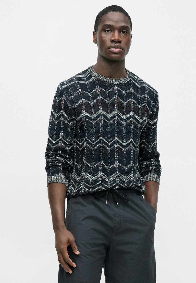 Pull avec un motif géométrique en chevron bleu foncé, gris et rouge, doté d'un tricot texturé et d'un col en côtes, associé à un pantalon sombre.