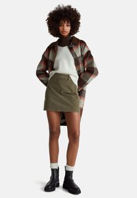 Karierter Wollmantel in Braun, Rost und Creme; weißer gerippter Pullover; olivgrüner Rock; schwarze Stiefeletten; cremefarbene gerippte Socken.