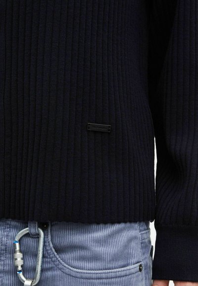 Pull côtelé bleu marine avec une petite étiquette de marque noire, assorti à un pantalon en velours côtelé bleu clair avec un mousqueton en métal accroché à la passants de ceinture.