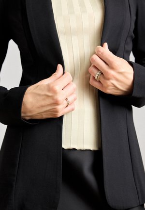 Blazer nero con una vestibilità sartoriale, indossato sopra un top beige a coste. Le mani si chiudono attorno al blazer, mostrando un anello d'argento e un anello di dichiarazione dorato.