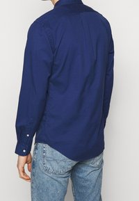 Tmavě modrá košile s dlouhým rukávem, límečkem na knoflíky, hladkou texturou a střihovou úpravou. Má rozdělený zadní díl a zapínání na knoflíky u zápěstí.