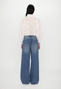 Chemisier semi-transparent blanc avec texture florale et manches bouffantes, associé à un jean en denim bleu à jambe large. Design simple, sans motifs visibles.