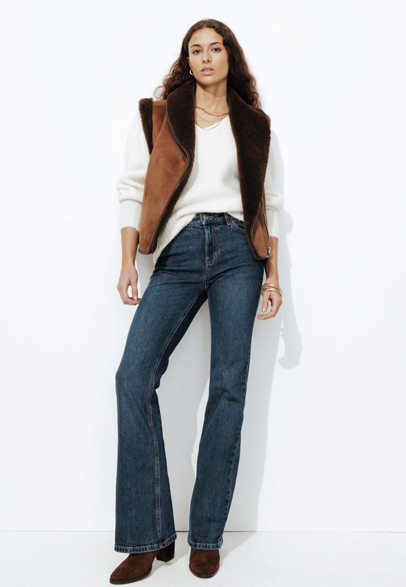 Gilet en daim marron avec un col en peluche, pull blanc, jeans évasés bleu foncé et bottines marron. Accompagné de bijoux simples.