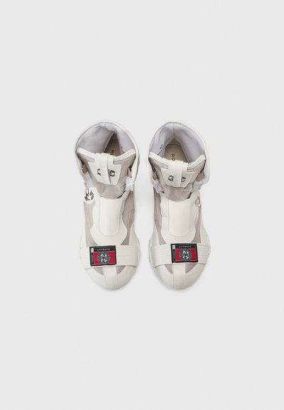 Baskets montantes à lacets en maille beige et blanche, avec un patch logo noir et rouge, des sangles élastiques et un design de bout arrondi.
