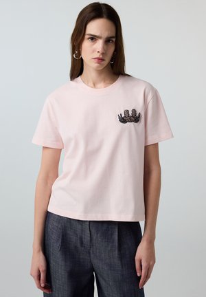 Joven mujer que lleva una camiseta de manga corta de color rosa claro con un diseño pequeño de araña negra y rosa en el pecho izquierdo y pantalones oscuros.
