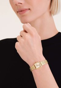 Reloj rectangular dorado con correa de eslabones metálicos y una carátula minimalista. Una mano sostiene un dedo cerca de la carátula, mostrando el reloj.