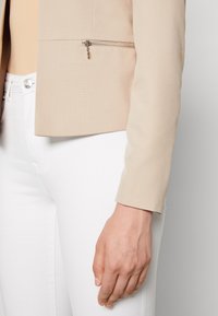 Veste ajustée beige avec une texture lisse, dotée d'une poche zippée sur le côté gauche, portée sur un pantalon blanc. Détails de design épurés et modernes.