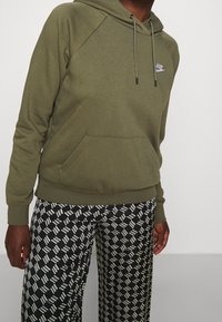 Zielona oliwkowa bluza z kapturem, ze sznurkami, kieszenią kangurka i ściągaczami na rękawach, zestawiona z szerokimi spodniami w czarno-biało-niebieski wzór.