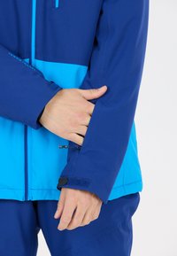 Blå och aqua jacka med texturerad yta. Har justerbara ärmslut med kardborre och en ficka med dragkedja. Modern, sportig design.