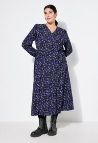 Lungo vestito floreale viola con motivi bianchi e blu, con scollatura a V, maniche lunghe e vita arricciata, abbinato a stivali neri.