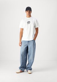 T-shirt en coton blanc avec le texte "LAND CRUISER". Jean en denim bleu clair à coupe décontractée. Casquette noire. Baskets blanches avec des rayures rouges. Fond uni.
