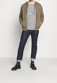 Sudadera verde con cremallera, camiseta de rayas blancas y negras, vaqueros oscuros de mezclilla con puños remangados, botines de cuero negro con detalle de costuras.